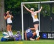 Field hockey players celebrate a goal as the goalkeeper grapples with the ball on the turf field during a match.