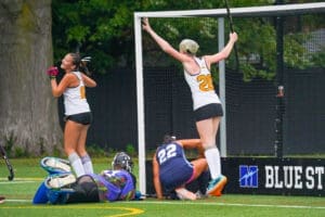 Field hockey players celebrate a goal as the goalkeeper grapples with the ball on the turf field during a match.