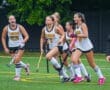 A group of excited female field hockey players in uniforms celebrates on the field after scoring a goal.