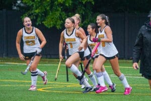 A group of excited female field hockey players in uniforms celebrates on the field after scoring a goal.