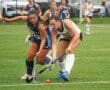 Two female field hockey players compete for the ball on a rain-soaked turf field during an intense match.