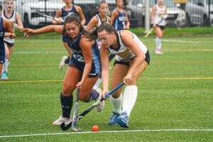 Two female field hockey players compete for the ball on a rain-soaked turf field during an intense match.