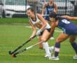 Two field hockey players compete for the ball on a wet turf field, showcasing athleticism and teamwork during the match.