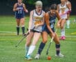 Field hockey players compete for the ball during a rainy game on a turf field, showcasing teamwork and athleticism.