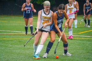 Field hockey players compete for the ball during a rainy game on a turf field, showcasing teamwork and athleticism.