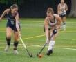 Two young female field hockey players competing for the ball on a wet turf field, showcasing athleticism and teamwork.