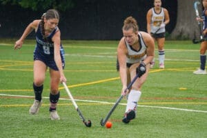 Two young female field hockey players competing for the ball on a wet turf field, showcasing athleticism and teamwork.
