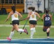 Three female field hockey players sprinting on a wet turf field, wearing uniforms and holding sticks, portraying competitive action.