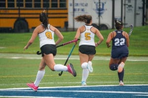 Three female field hockey players sprinting on a wet turf field, wearing uniforms and holding sticks, portraying competitive action.