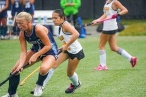 Two young athletes compete in a field hockey match, showcasing skill and determination on a rainy day.