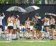 Field hockey players take shelter under umbrellas during a rainy practice, showcasing teamwork and resilience.