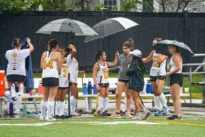 Field hockey players take shelter under umbrellas during a rainy practice, showcasing teamwork and resilience.