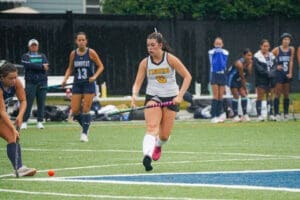 High school field hockey player in action during a rainy game, showcasing skills on the turf with teammates in the background.