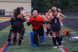 High school field hockey team celebrates a goalie in orange uniform running through a cheering line of teammates.