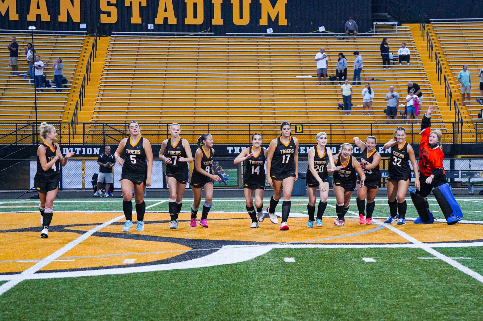 High school soccer team in black uniforms celebrating on the field at Herman Stadium, with cheering fans in the stands.