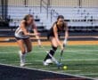 Two female field hockey players compete for the ball on a turf field during a match, showcasing teamwork and sportsmanship.