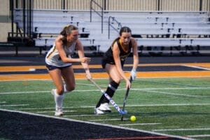 Two female field hockey players compete for the ball on a turf field during a match, showcasing teamwork and sportsmanship.
