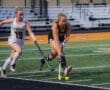 Two female field hockey players compete for the ball on a brightly colored turf field during an evening match.