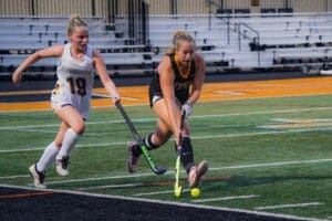 Two female field hockey players compete for the ball on a brightly colored turf field during an evening match.