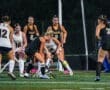 High school field hockey match in progress, players in action with lights illuminating the field at night.