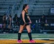 Field hockey player in action, wearing a black jersey and bright pink shoes, focused during a rain-soaked game.