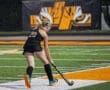 Field hockey player in action on the turf, focusing on the ball during a night game with vibrant team colors in the background.