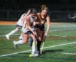 Two field hockey players compete for the ball under the night lights on a turf field. Intense action during the game.