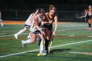 Two field hockey players compete for the ball under the night lights on a turf field. Intense action during the game.