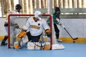 A goalie in a white Tigers jersey defends the net while players compete in a roller hockey game.
