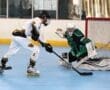 Roller hockey players compete in an intense game, with a player shooting on goal while the goalie prepares for the save.