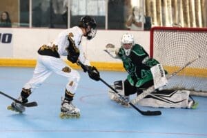 Roller hockey players compete in an intense game, with a player shooting on goal while the goalie prepares for the save.