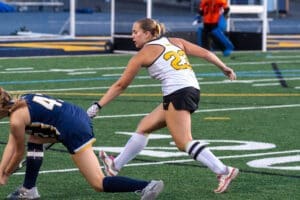 Field hockey players in action, with one athlete wearing jersey number 23 evading a defender on the turf.