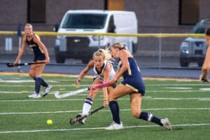 Two female field hockey players compete for the ball on a synthetic turf field during an evening match.