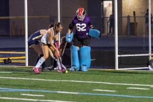 Field hockey players in action; a goalie prepares as a teammate approaches the ball near the goalpost.