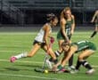 Field hockey players compete for the ball during an intense game on a well-lit turf field at night.