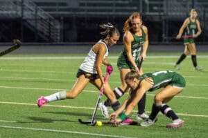 Field hockey players compete for the ball during an intense game on a well-lit turf field at night.