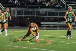 High school field hockey players compete under stadium lights; a player lunges to hit the ball on the field.