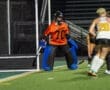Goalie in an orange jersey prepares to defend the net during a field hockey match under stadium lights.