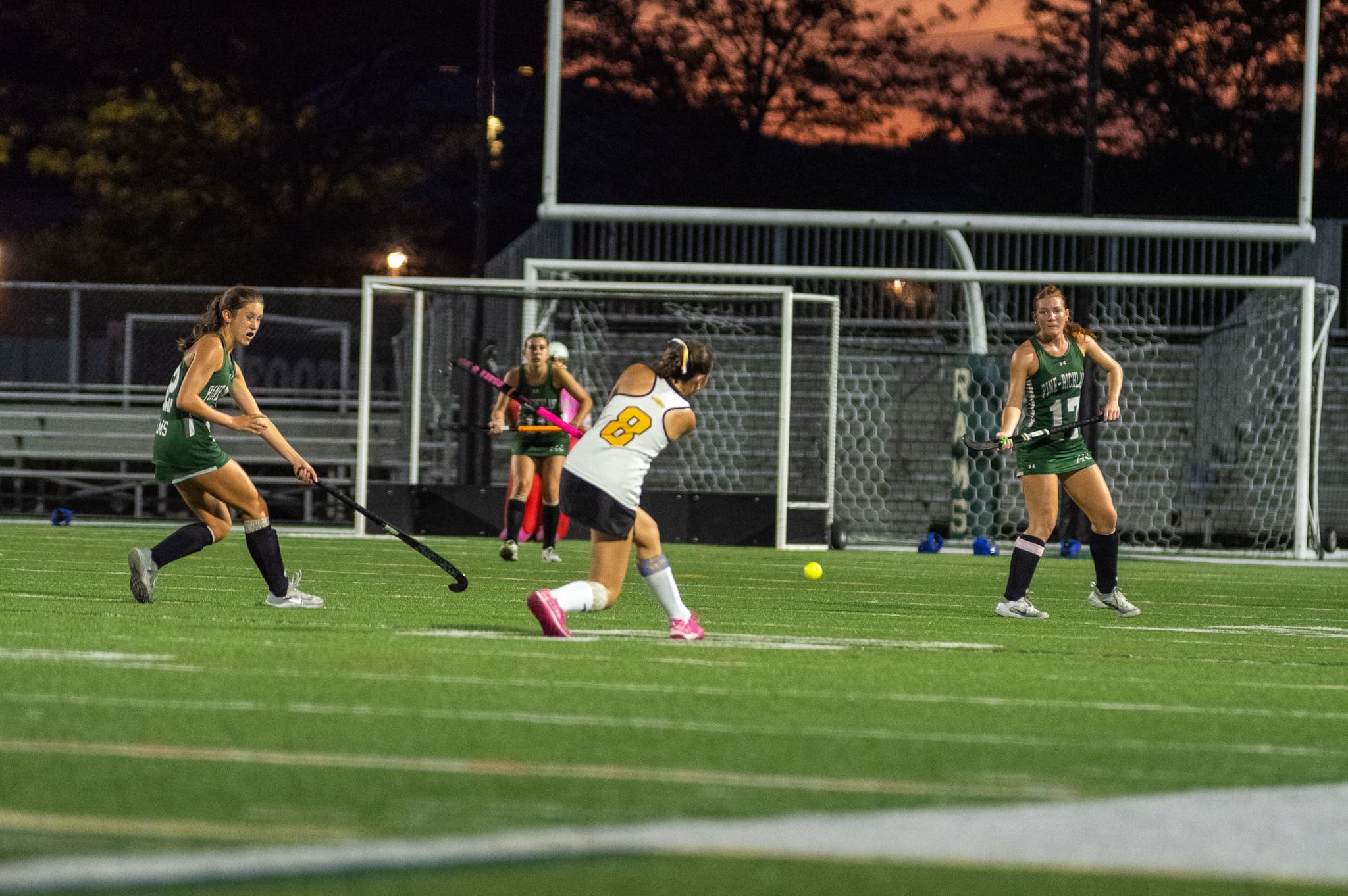 Field hockey players in action during an evening match on a stadium field, showcasing dynamic gameplay and teamwork.