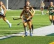High school field hockey players compete intensely, with one athlete dribbling the ball while teammates pursue.