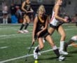 High school field hockey player dribbling the ball during a competitive match on a turf field at night.