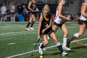 High school field hockey player dribbling the ball during a competitive match on a turf field at night.