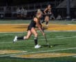 High school field hockey player dribbles the ball during a night game on an artificial turf field. Team in the background.