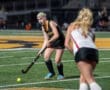 Field hockey player prepares to strike the ball on a turf field during an intense game. Team jerseys visible in background.