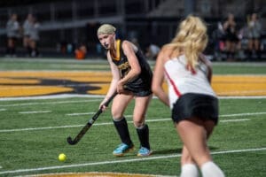 Field hockey player prepares to strike the ball on a turf field during an intense game. Team jerseys visible in background.