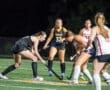 Two field hockey players compete for the ball under the lights, with teammates watching on the field.