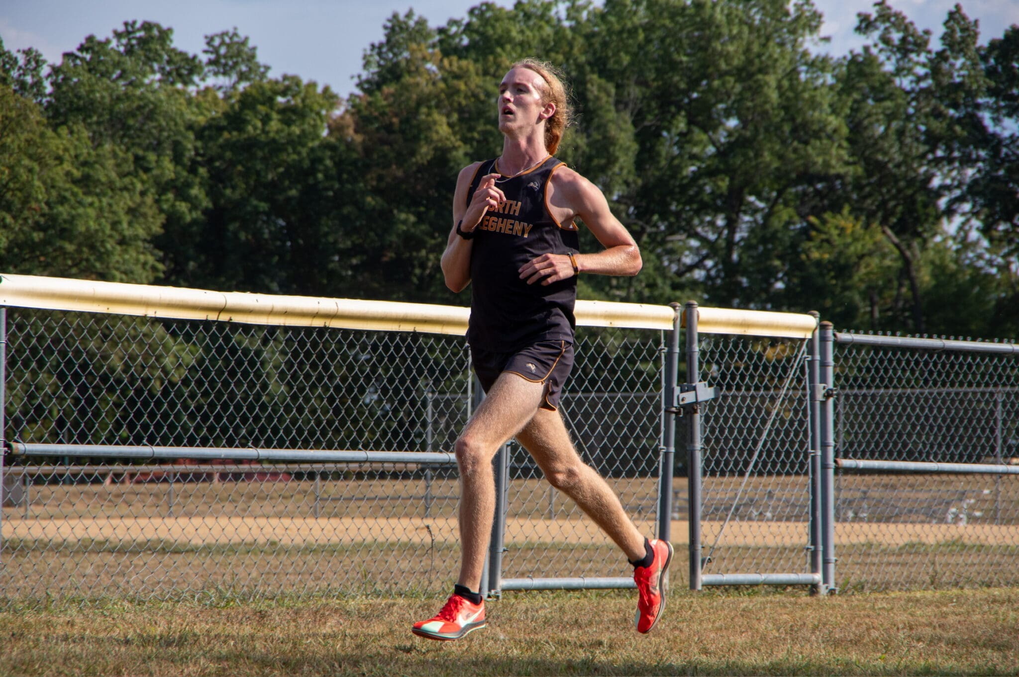 Athlete running on a field, wearing a black jersey and red shoes, with trees and a fence in the background.
