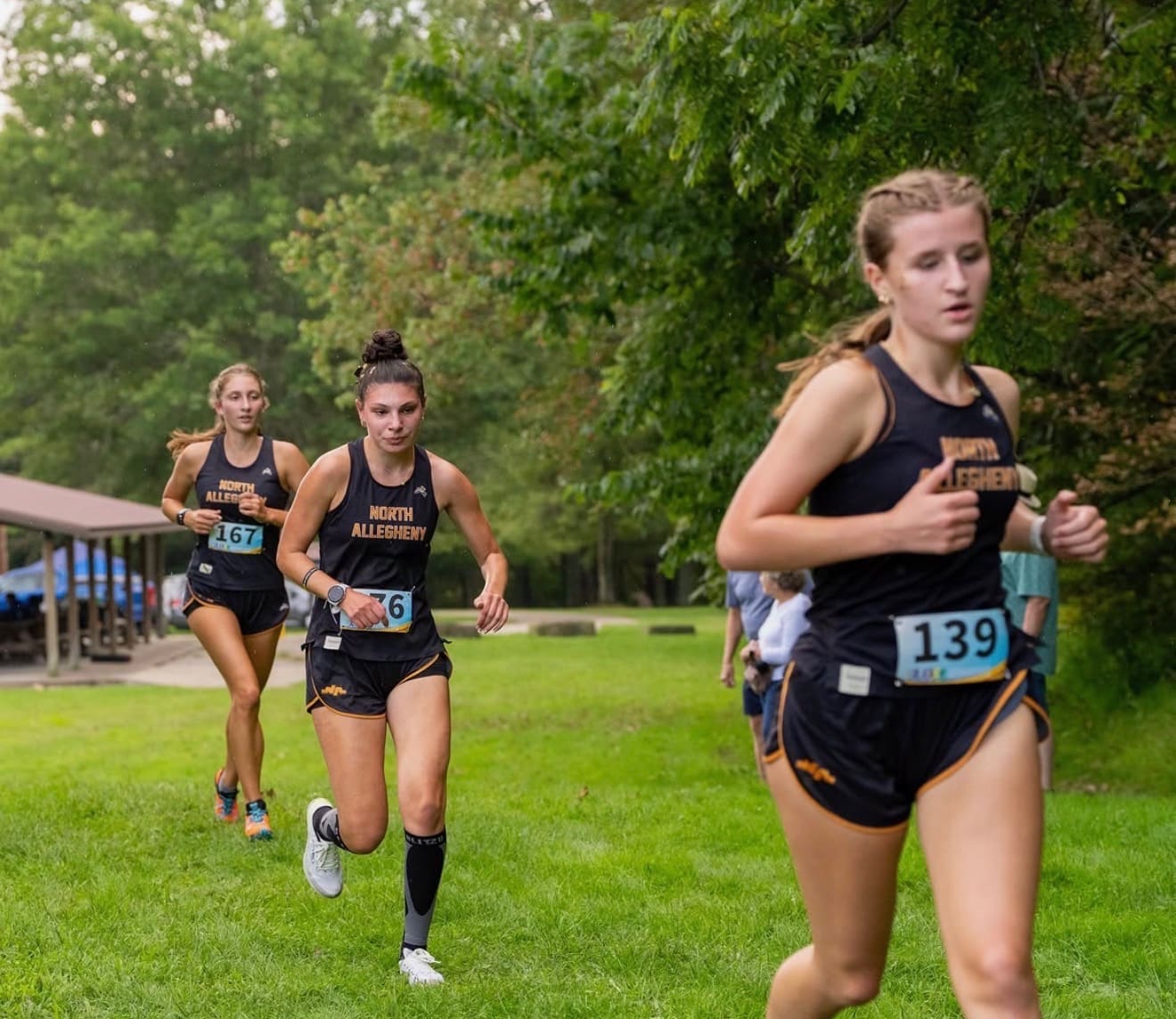 Three female cross country runners from North Allegheny sprinting on a grassy trail during a race.