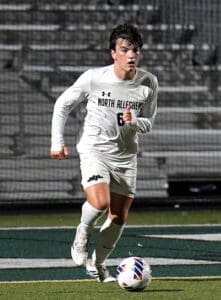 Player in white uniform of North Allegheny runs with the soccer ball during a night game on the field.