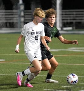 Two high school soccer players compete for the ball on a field at night, with action-packed intensity.